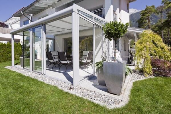 Moderner Wintergarten aus Glas mit Gartenmöbeln an Einfamilienhaus im Grünen.